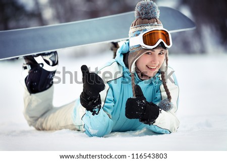 Young beautiful female snowboarder resting on ski slope, she\'s lying on front, showing thumbs up and smiling, close up