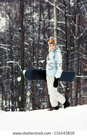 Young pretty woman holding her snowboard and walking on ski slope in sunlight, wood on background, side view, copy space