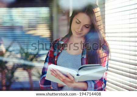 Young woman with a book standing near window; shot through window glass with reflections and flares