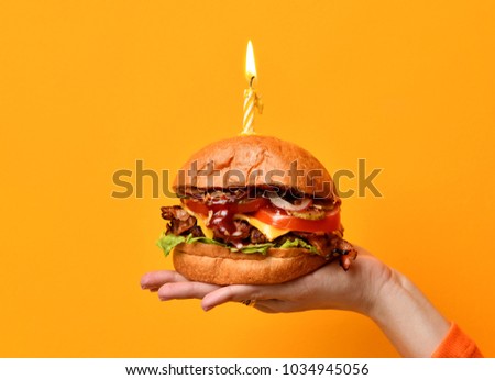 Woman hands hold big burger barbeque sandwich with beef and lit candle for birthday party on yellow background