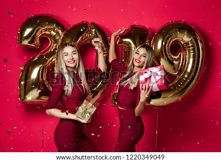 Two women celebrating at new year party happy laughing girls in casual dresses throw gold stars confetti having fun with 2019 balloons on red background