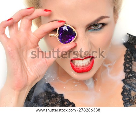 Portrait of angry fashion sexy woman face lying in milk bath with splashes red lips and nails holding gold pendant with big amethyst and diamonds on white background