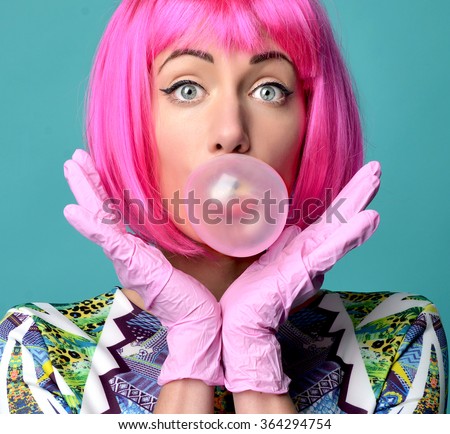 Close up funny fashion portrait of cheerful woman inflating the bubble gum in hot pink party wig on a mint background.