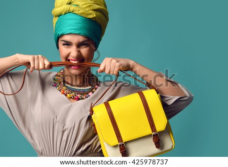 Woman yelling screaming and eating belt of hand hold stylish fashion yellow leather bag handbag isolated on blue mint background