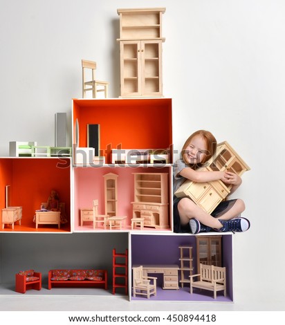 Red hair Baby Girl Kid playing with doll house stuffed with mini furniture toys and doll sitting on a cube in play room at home or kindergarten hugging little cabinet happy smiling