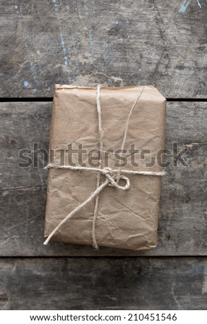 A small package wrapped in recycled brown paper which is tied with a white cord. The package is lying on weathered wood.