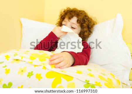 Curly haired little sick girl laying in bed and blowing her nose.
