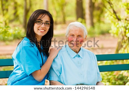Kind elderly lady with caring nurse sitting both on bench in a park.