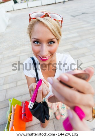 Cute blond girl with shopping bags holding lipstick and mirror from wide angle.
