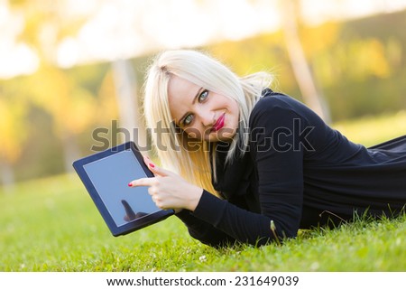 A young woman pointing towards a blank tablet outdoors on the grass.