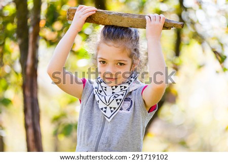 Playful little girl holding a wooden stick on her head outdoors in a park.