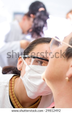 A young dentist examinig an old woman\'s teeth, doctors in the background - part of a series.