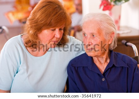An old woman next to a mature one, a mother and a daughter, the elderly woman is sitting on a wheelchair having a worried facial expression.
