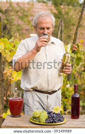 An elderly man holding a bottle and a glass of white wine in his hand, fresh grapes, a jug of must and a bottle of red wine in front of him on an old wooden table.
