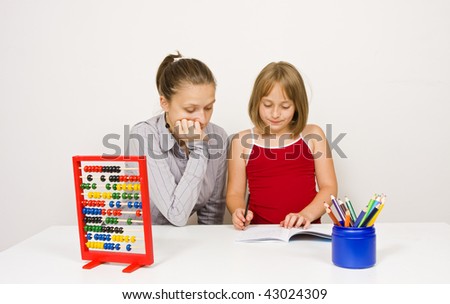 A teacher, or mother, learning together with her student, or daughter, paying attention to the little girl\'s action agains white wall - learning tools and educational objects on table.