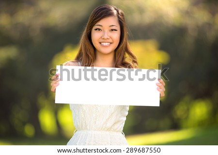 A beautiful young adult woman holding a blank white sign card outdoors smiling confident