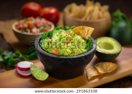 A delicious Bowl of Guacamole next to fresh ingredients on a table with tortilla chips and salsa