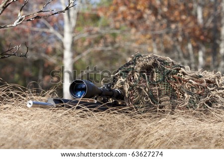 sniper laying on the ground covered in a ghille suite tall grass and trees in the background