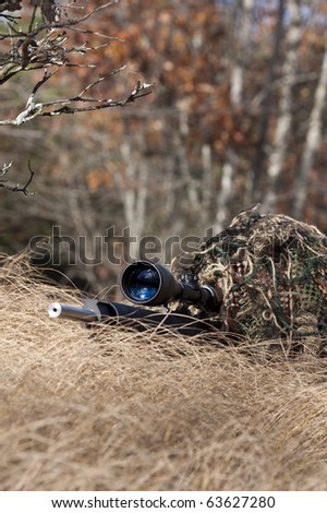 sniper laying on the ground covered in a ghille suite tall grass and trees in the background