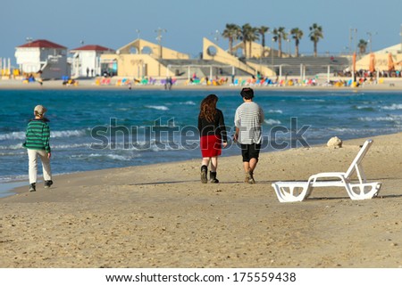 A young jewish cupple is walking on the beach in Natanya, Israel. A boy is walking behind them. On the background are two lifeguard towers and a caffe.