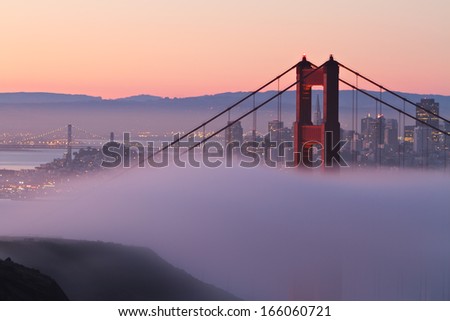 The north tower of the Golden Gate Bridge draped in morning fog with beautiful San Francisco in the background.
