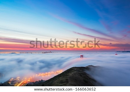 Fog pours into the bay and over the Golden Gate Bridge as sunrise lights the clouds over San Francisco, California.