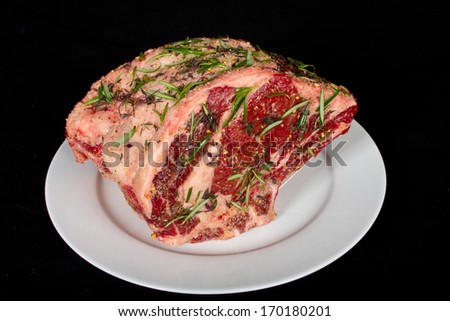 Close up of a beautiful delicious raw angus standing prime rib roast with rosemary and thyme herb seasoned on a white plate and ready to slow roast in the over.