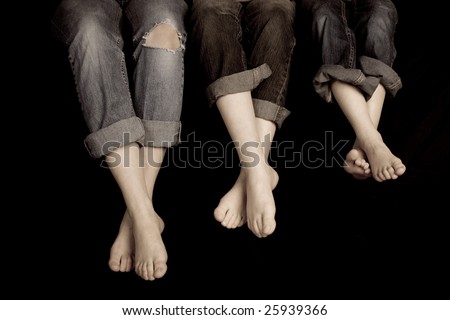 Three pairs of feet in jeans rolled up on a black background. One pair of jeans ripped at the knee\'s