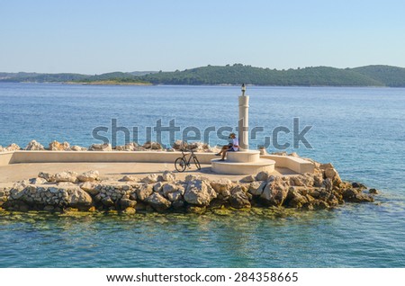 Anonymous lonely young man with a bike reading by the lighthouse on a stone pier by the blue sea