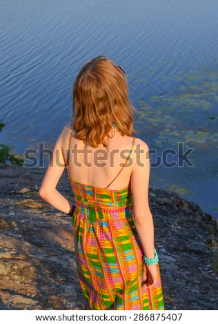 A young slim redhead girl in colorful summer dress with open sexy back standing on the rock by the river