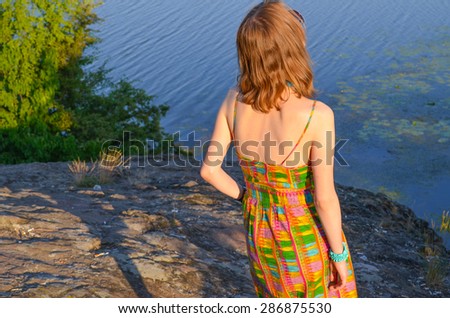 Attractive girl in summer dress standing on the rock by the river in sunlight. Back view