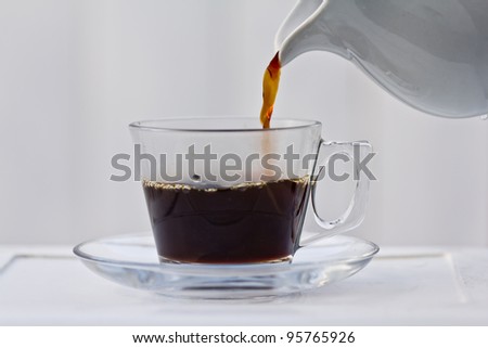 Pouring coffee into a half full clear cup with a saucer.