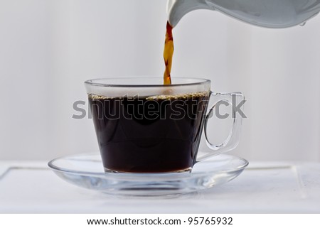 Pouring coffee into an almost full clear cup with a saucer.