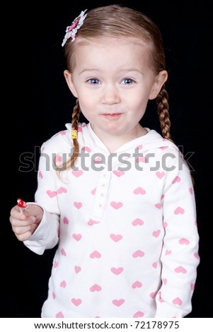An excited child eating a lolly pop.  She is wearing a white shirt with hearts on it.  She has an excited and funny look on her face.