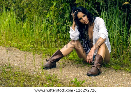 Sexy girl in denim shorts and white shirt sitting by the countryside road