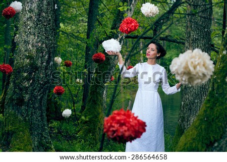Young beautiful woman in long white  dress walking in the forest with decorative flowers on the trees