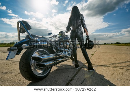 Biker girl in leather jacket standing by a motorcycle. Rear view