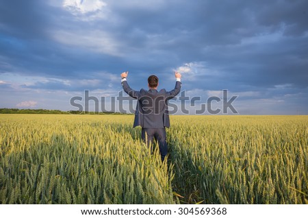 happy farmer, businessman, standing in wheat field over wind turbines background with his hands up and thumbs up