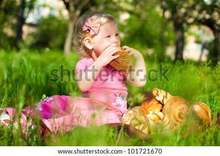 The little blonde beautiful girl sitting on a green lawn in the park and eat a muffin.