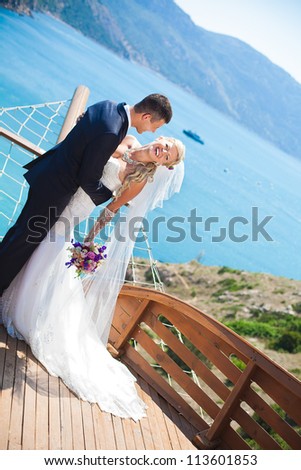 Couple in love bride and groom posing in a restaurant stylized as deck wooden ship on the background of the bay with beautiful cliffs. Enjoying a love and happiness on their wedding day in the summer.