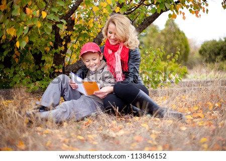 Beautiful family of mother and son posing on a background a tree with yellow leaves and grass gray. Healthy young mother with a luxurious blonde hair. Autumn.
