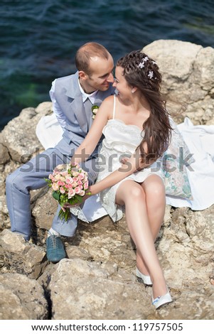 Happy pregnant bride and groom holding wedding bouquet posing against the sea. The groom tenderly embraces the pregnant belly of his wife.