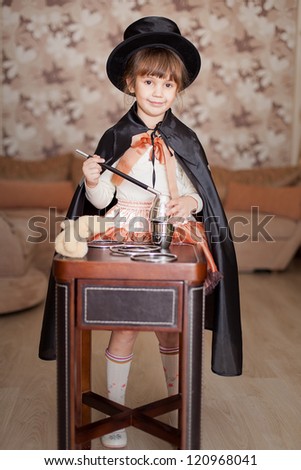 Cute child posing in Attire conjurer with a magic wand at the table with magical accessories at home. Series.