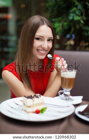 Attractive young woman has a rest in restaurant in front of her on the table tiramisu cake and drink coffee latte in a glass beaker.