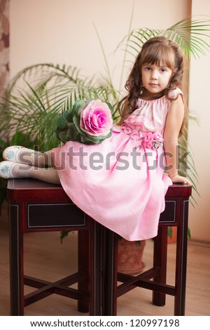 Beautiful little girl model view in a luxury pink dress and flower posing at home on the table.