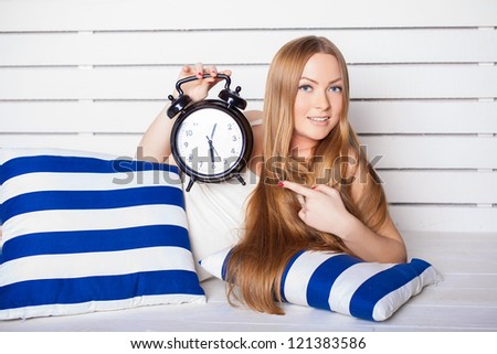 Beautiful young woman lying on the bed. In the foreground is a big clock. Morning awakening