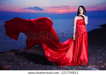 Beautiful woman posing in a luxurious red dress with a long train standing at the cliff against the sea.