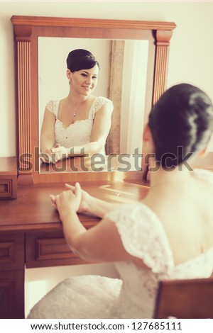 wedding: Happy beautiful brunette bride sitting on chair and looking through mirror