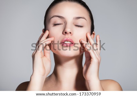 Naturally beautiful brunette woman with flawless skin touching her face dreamily over gray background