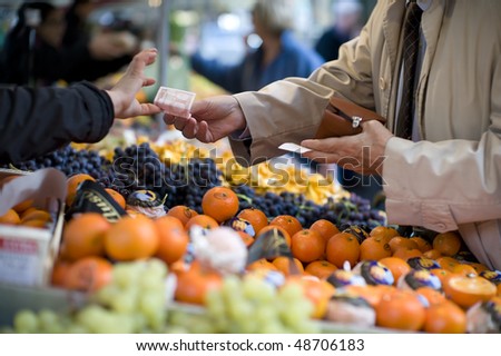 A man reaches across a row of fruit in an outdoor market to pay a vendor with a 10 Euro bill. Focus is tight on the bill changing changing hands. The foreground and background are out of focus.